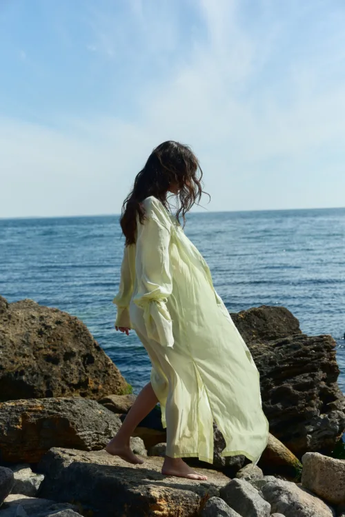 Person walking barefoot on rocky shoreline in a pale yellow dress with the sea behind them.