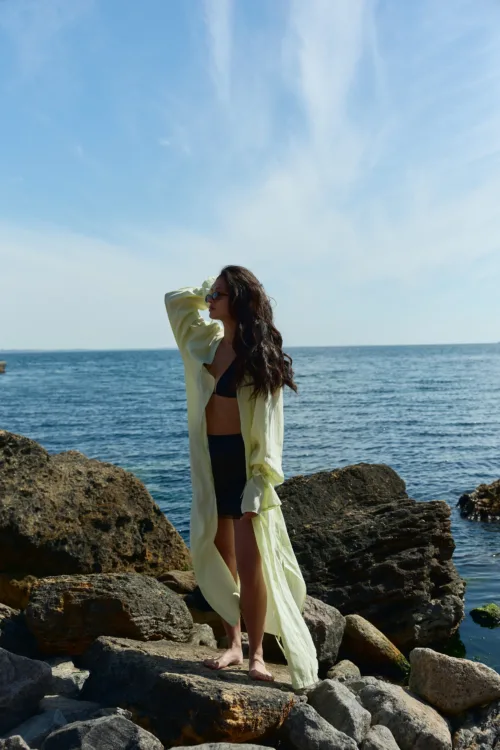 Woman in a black bikini with a pale yellow cover-up standing barefoot on rocky shoreline, ocean and blue sky behind her.
