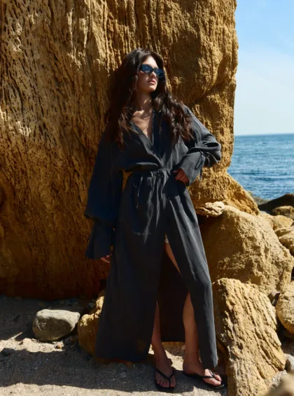 Woman in a dark flowing dress leans against a large textured rock cliff at the beach, wearing sunglasses, with the sea in the background.