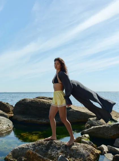 Young woman in a bikini top and yellow shorts walks barefoot on rocky shore with a dark cape billowing behind her against a blue sky and ocean backdrop.