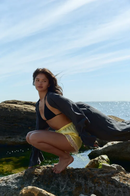 Woman crouching on sunlit rocky shoreline by the sea, wearing a black bikini top and yellow shorts, with a gray cover-up flowing behind her.