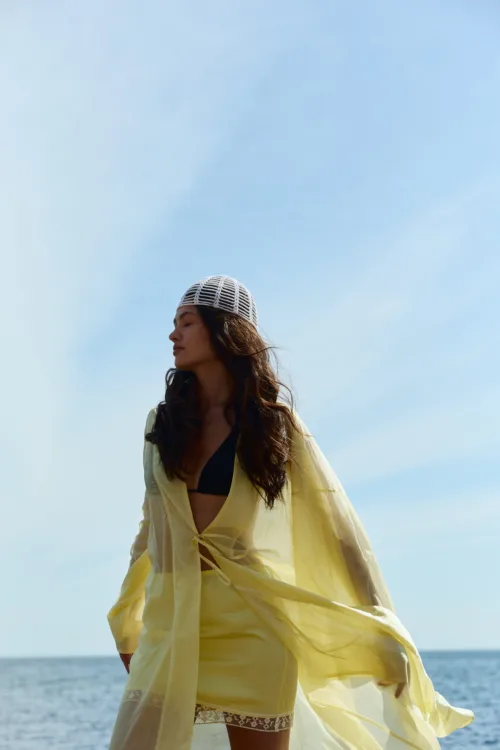 Woman standing at the beach in a yellow sheer cover-up over a black bikini, with a white knitted cap, against a blue sky and ocean.