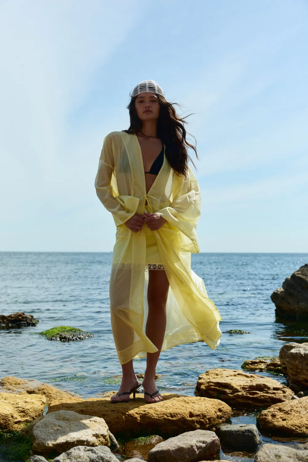 Woman standing on rocky shore in a yellow sheer cover-up over a black bikini, with the sea and blue sky behind her and a white knit cap on her head.