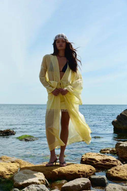 Woman standing on rocky shore in a yellow sheer cover-up over a black bikini, with the sea and blue sky behind her and a white knit cap on her head.
