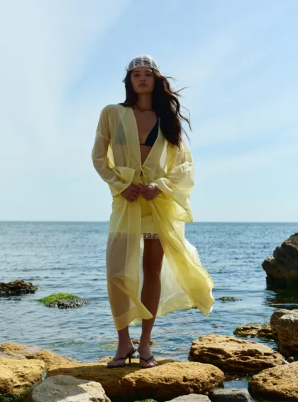 Woman standing on rocky shore in a yellow sheer cover-up over a black bikini, with the sea and blue sky behind her and a white knit cap on her head.