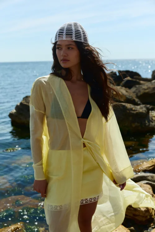 Woman in a pale yellow sheer cover-up over a black bikini, standing by the rocky shoreline with the ocean in the background and a white woven hat on her head.