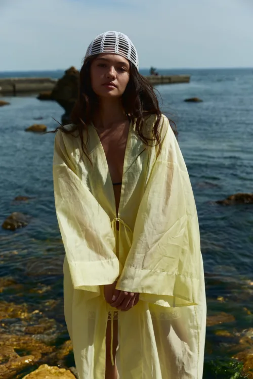 Woman standing by a rocky shoreline, wearing a pale yellow cover-up and a white knit cap, with the sea in the background.
