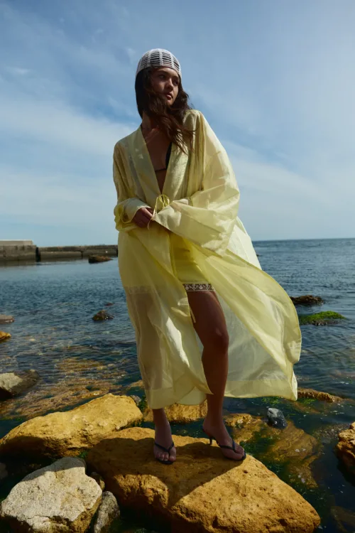 Woman standing on rocky shore in a pale yellow cover-up, by the sea under a blue sky, wearing a hat and flip-flops.