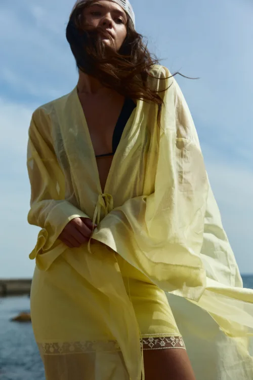 Woman in a pale yellow sheer dress, breeze whipping her hair, standing outdoors by the water. Notable lace trim at the hem.