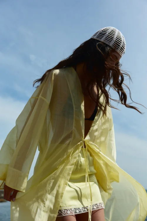 Person wearing a pale yellow sheer dress and white knit hat, with wind-blown hair against a blue sky.