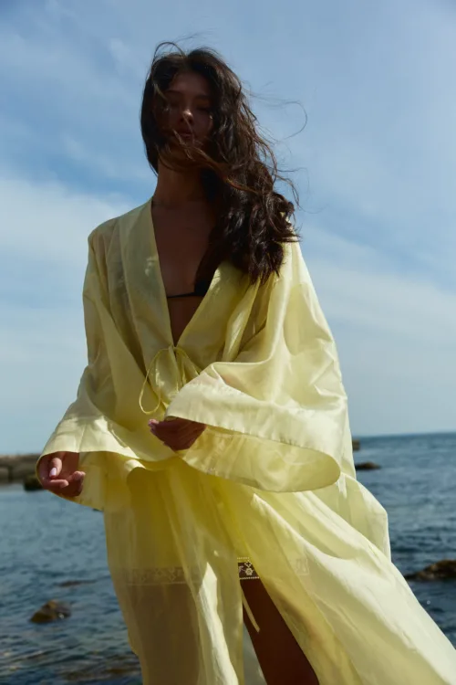 Woman at the beach wearing a light yellow sheer cover-up over a black bikini, hair blown by the wind with blue sky and sea in the background.
