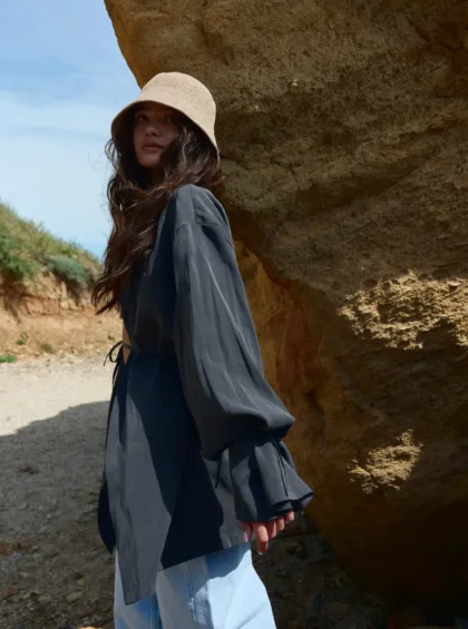 Young woman wearing a beige bucket hat and dark jacket stands next to a large rock formation outdoors, sunny day.