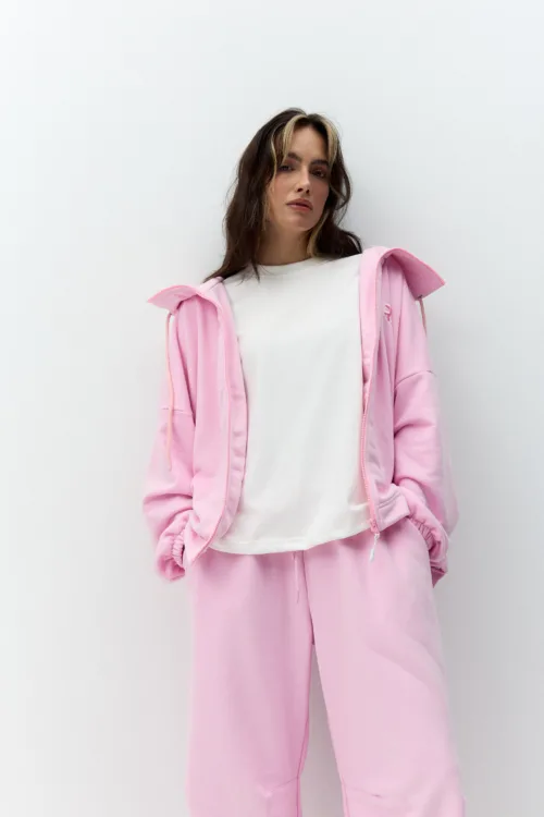 Woman wearing a pale pink hoodie and matching sweatpants over a white T‑shirt, posing against a white wall with hands in pockets.",