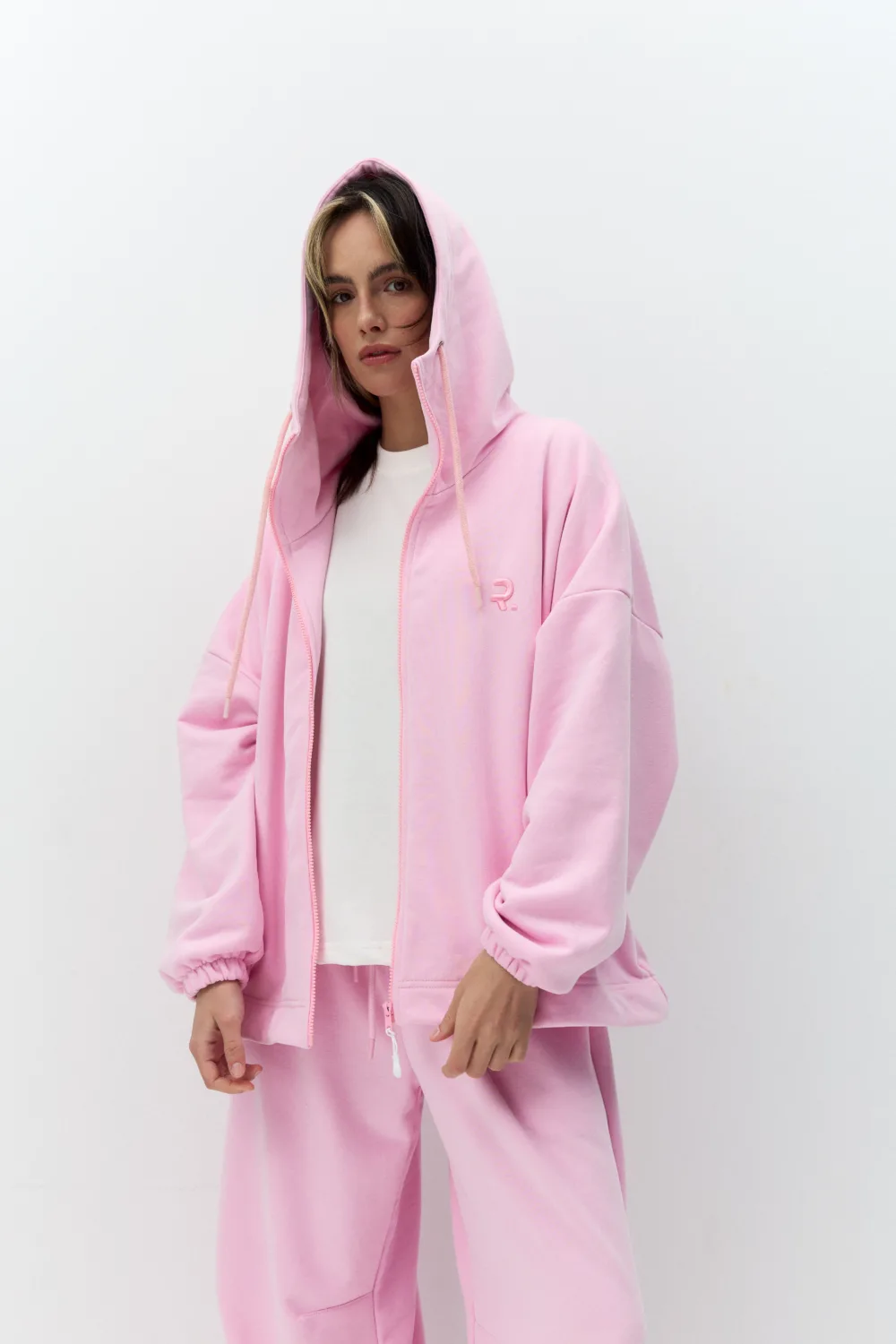 Person wearing a pastel pink hoodie and pants, white shirt underneath, standing against a white background, looking at the camera.