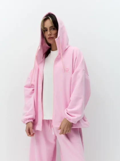 Person wearing a pastel pink hoodie and pants, white shirt underneath, standing against a white background, looking at the camera.