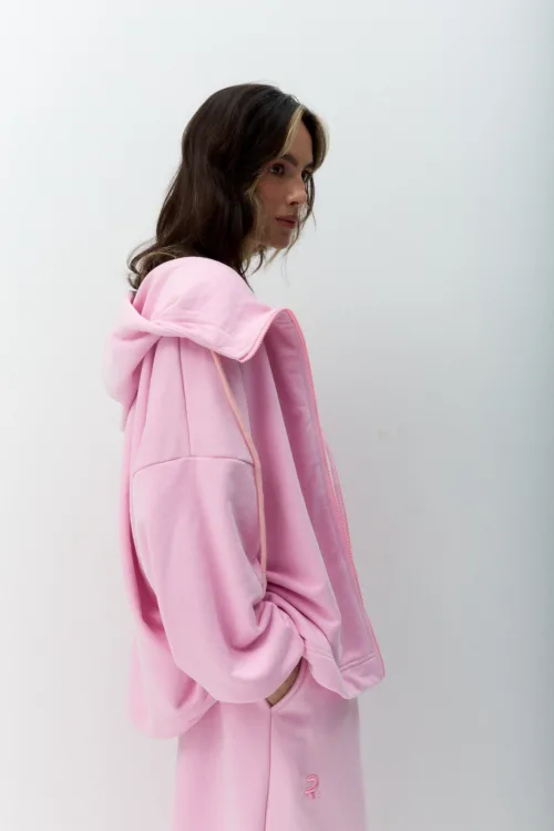 Side profile of a woman wearing a pink hoodie, hands in pockets, against a plain light background.