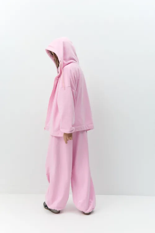 Side view of a person in a pink hoodie with the hood up, standing against a plain white wall.