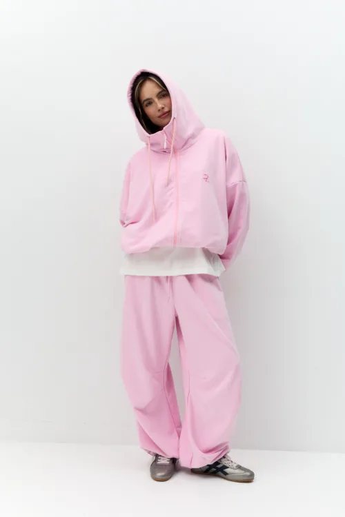 Person in a light pink hoodie and matching sweatpants standing against a plain white backdrop, hands in pockets and hood up.