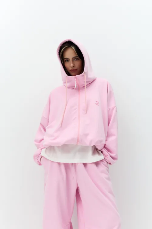 Woman with dark hair wearing a pink hooded sweatshirt, hood up, standing with hands in pockets against a white wall.