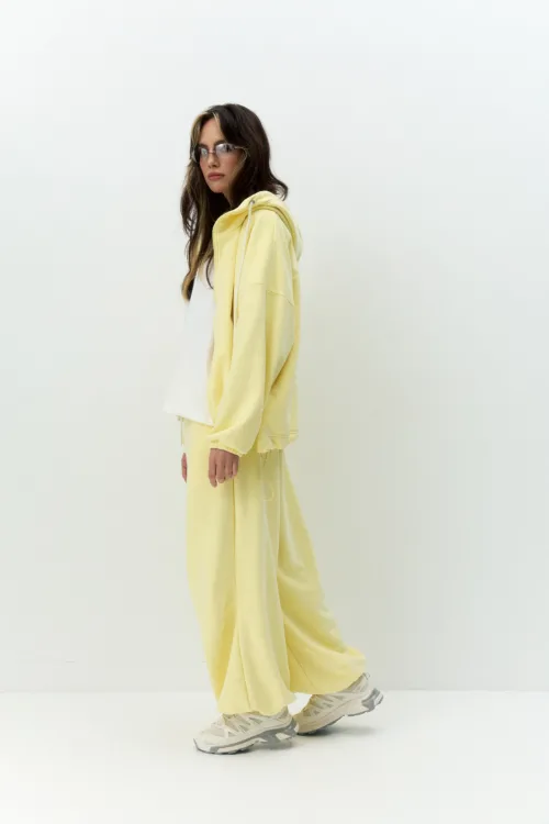 Side view of a person wearing a pale yellow hoodie and matching pants with white sneakers, standing against a plain light backdrop.