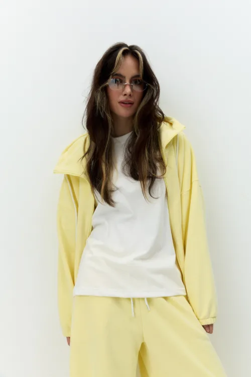 Woman in a pale-yellow hoodie and matching joggers over a white T‑shirt, wearing tinted sunglasses, posed against a light wall.