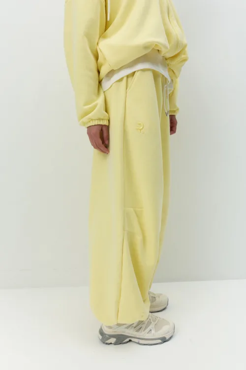 Person in pale yellow tracksuit, hoodie, and sneakers, standing with one hand at side (side view).