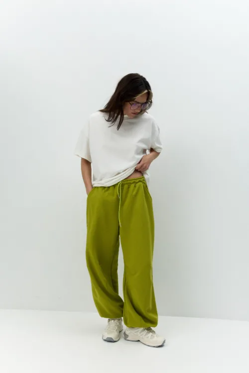 Person standing against a plain white wall, wearing an oversized white T-shirt, olive-green jogger pants, and sneakers, looking down.