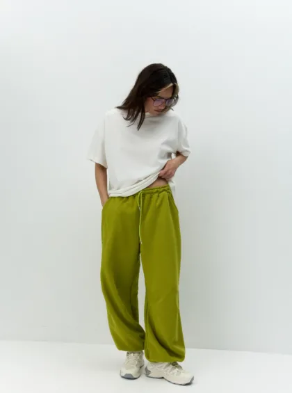 Person standing against a plain white wall, wearing an oversized white T-shirt, olive-green jogger pants, and sneakers, looking down.