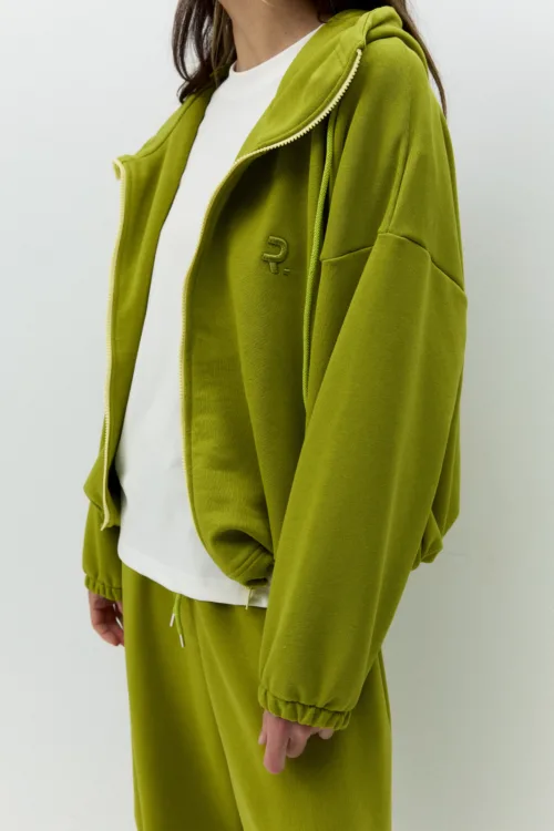 Person wearing an olive-green zip-up hoodie and matching sweats over a white T-shirt, with a subtle logo on the chest.