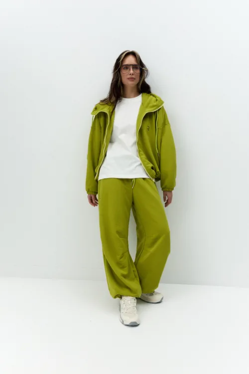 Person wearing a lime green hoodie and pants with a white T‑shirt and sunglasses, posed against a plain white wall.