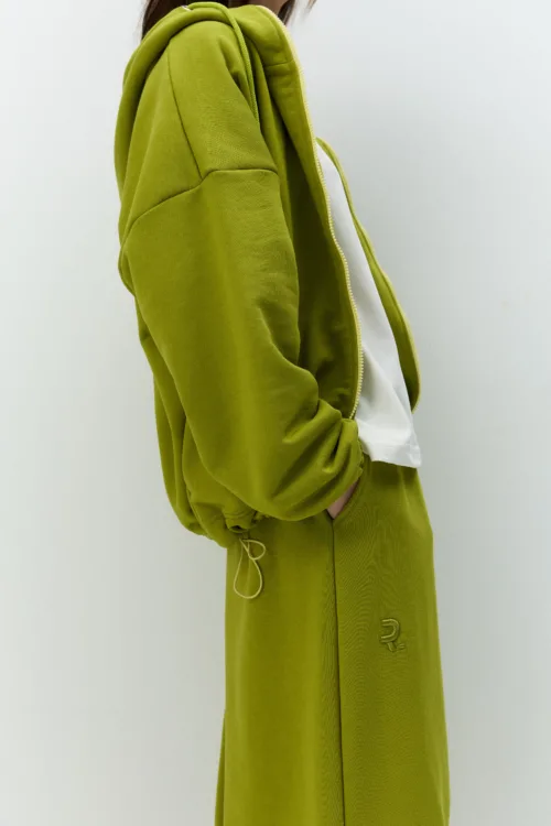 Person in an olive-green hoodie with a white shirt, standing in profile with hands in pockets.