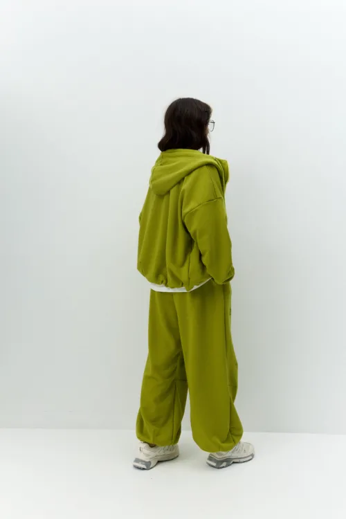 Olive-green hoodie and sweatpants worn by a person seen from behind, short dark hair and glasses, standing against a plain white wall.