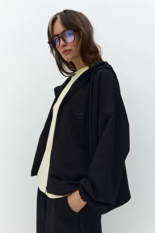 Woman in a black coat over a cream top, wearing blue-tinted oversized sunglasses, standing against a plain pale background.