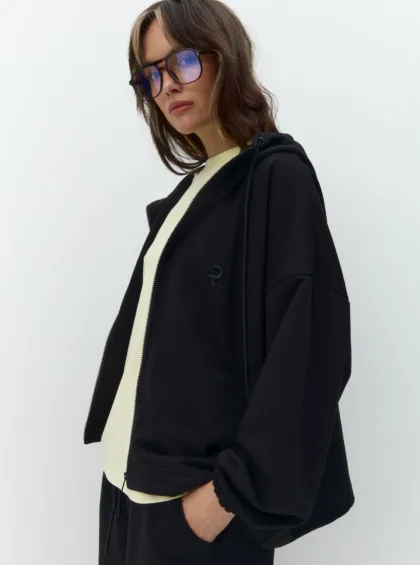 Woman in a black coat over a cream top, wearing blue-tinted oversized sunglasses, standing against a plain pale background.