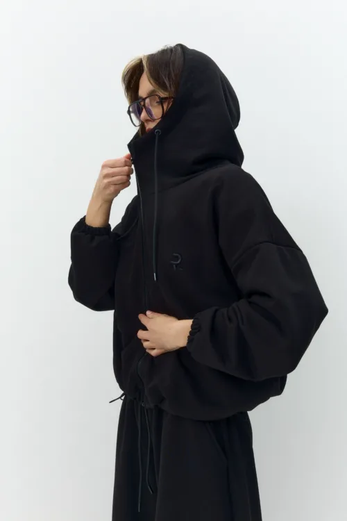 Person in a black hooded sweatshirt adjusting the zipper, facing left against a light background, with glasses on.