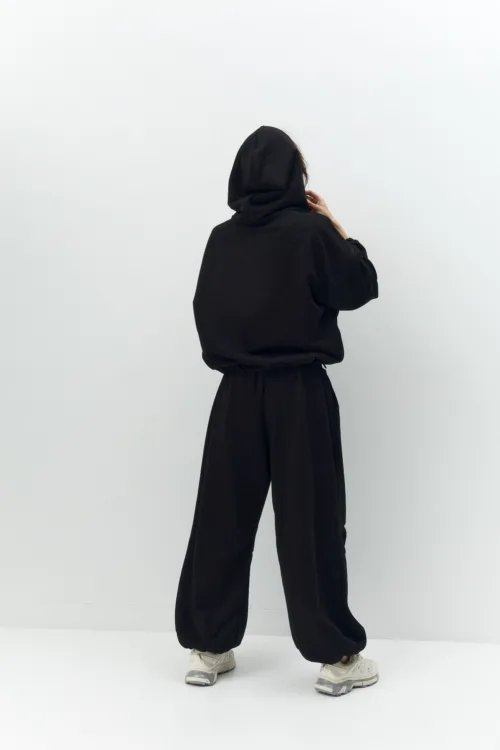 Person seen from behind, wearing a black hoodie and matching black pants, standing against a plain white wall.