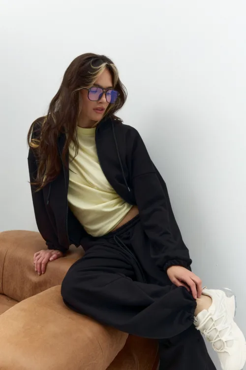 Woman sitting sideways on a tan sofa in a black jacket and pale yellow top, wearing blue-tinted sunglasses.