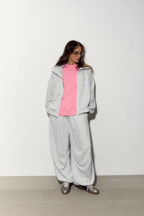 Woman in a pink t-shirt under a gray hoodie/joggers, sunglasses, standing against a white wall with hands in pockets.