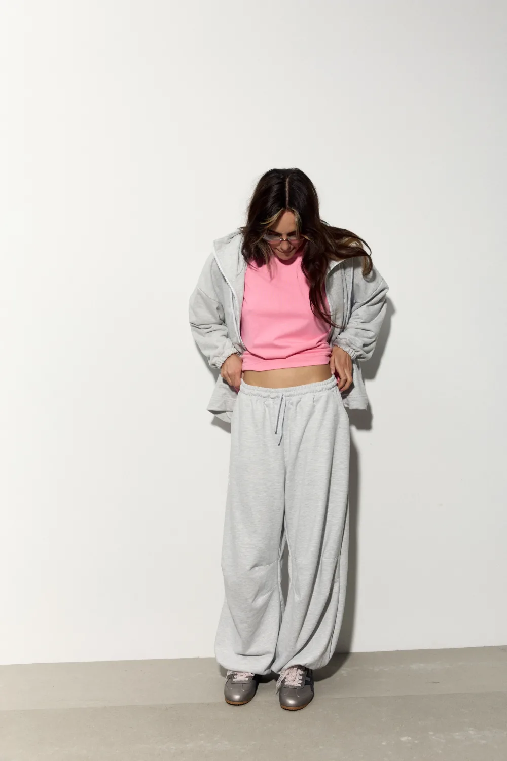 Person standing against a plain white wall, wearing a pink top under a light gray hoodie and sweatpants, looking down at their midsection.