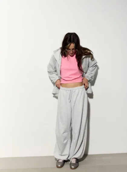 Person standing against a plain white wall, wearing a pink top under a light gray hoodie and sweatpants, looking down at their midsection.