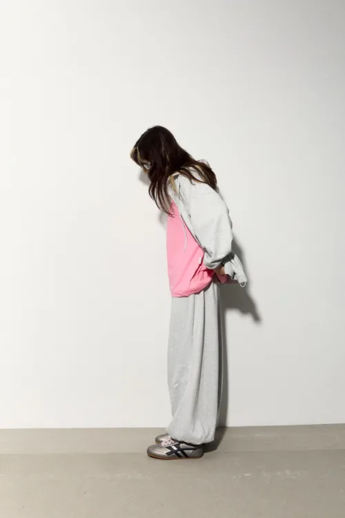 Person standing in profile, head lowered, wearing a gray hoodie over a pink top and light gray sweatpants with sneakers, against a plain white wall.