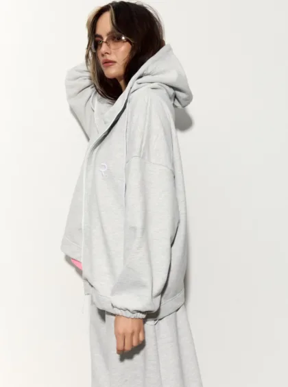 Person in a light gray oversized hoodie, shown in profile, wearing tinted sunglasses against a white background.