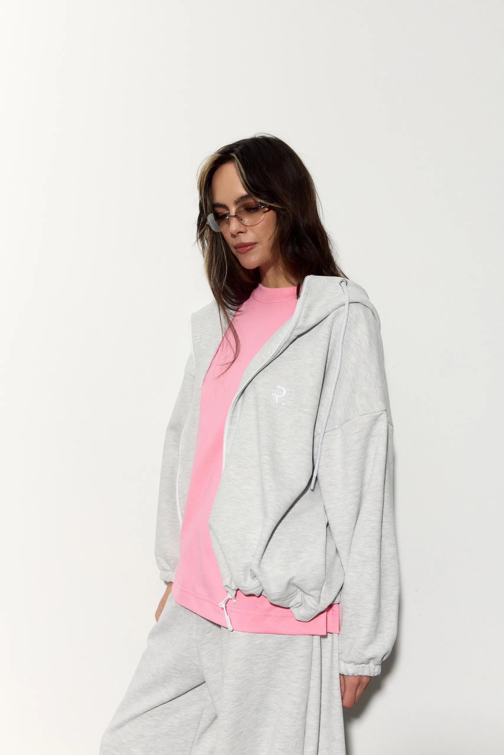 Жіноче oversized худі сірого кольору з футеру діагональ преміум. Вільний силует, блискавка та м’яка фактура для casual образів. - romanova4939_Original Худі жіноче oversized сіре на блискавці без утеплення - Woman in a pink shirt under a light gray zip hoodie, wearing sunglasses, posing against a white background.