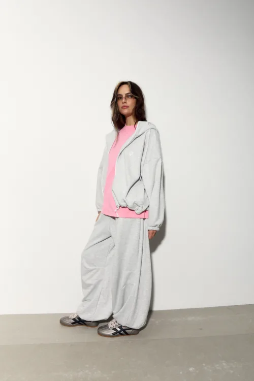 Woman in a pink top and gray oversized tracksuit, sunglasses, standing against a plain white wall in a fashion pose.