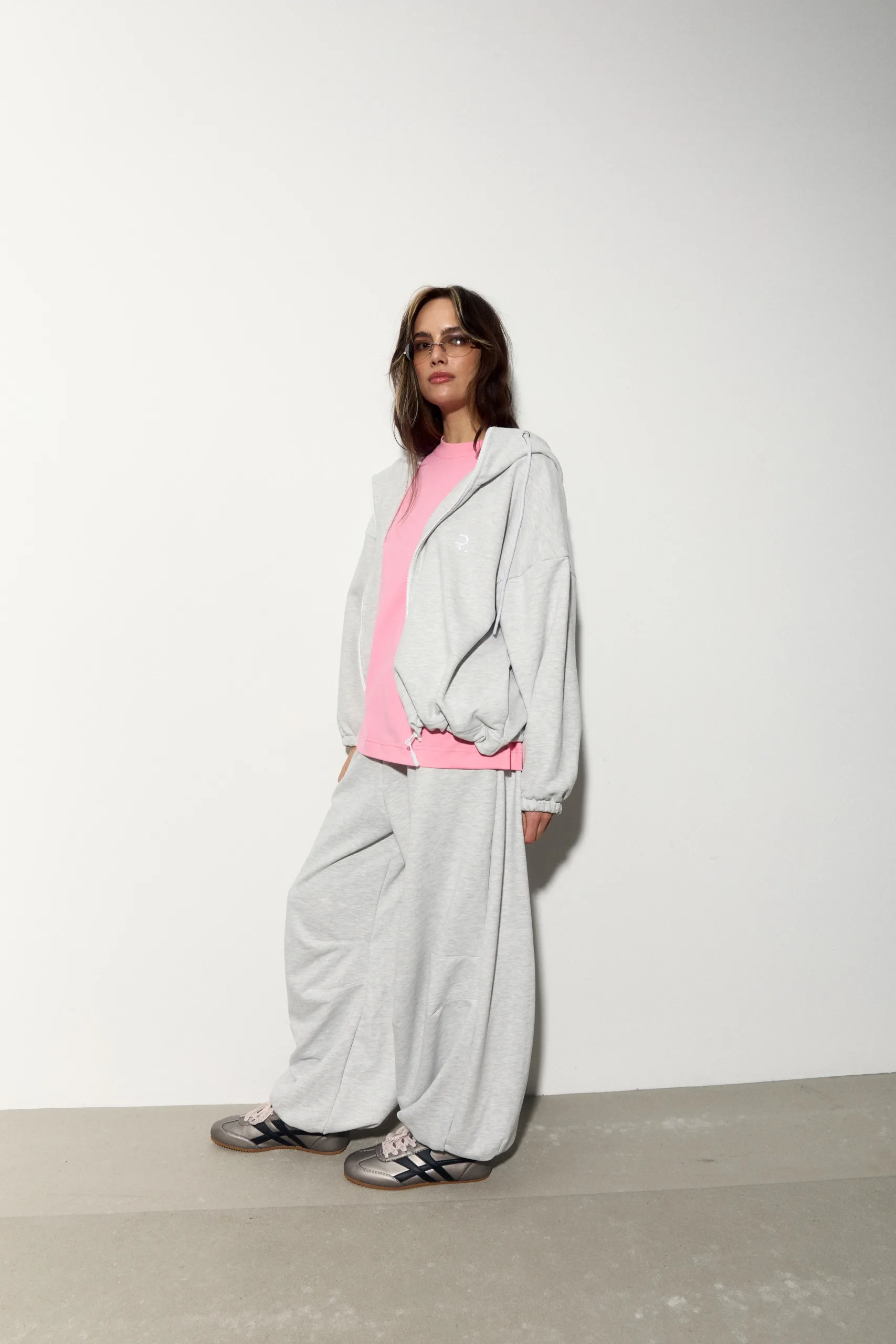 Жіноче oversized худі сірого кольору з футеру діагональ преміум. Вільний силует, блискавка та м’яка фактура для casual образів. - romanova4940_Original Худі жіноче oversized сіре на блискавці без утеплення - Woman in a pink top and gray oversized tracksuit, sunglasses, standing against a plain white wall in a fashion pose.