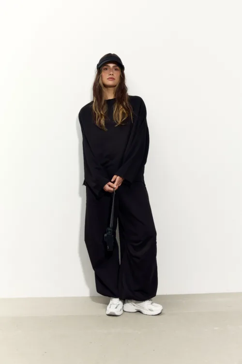 Young woman in a black oversized outfit and cap stands against a plain white wall, hands clasped at the waist, holding a bag strap.