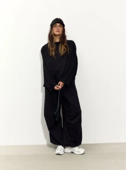 Young woman in a black oversized outfit and cap stands against a plain white wall, hands clasped at the waist, holding a bag strap.