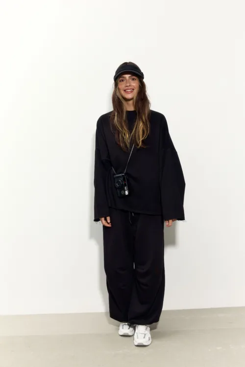Woman in an oversized black outfit with a black cap, smiling, and a camera hanging from her neck against a plain white wall.