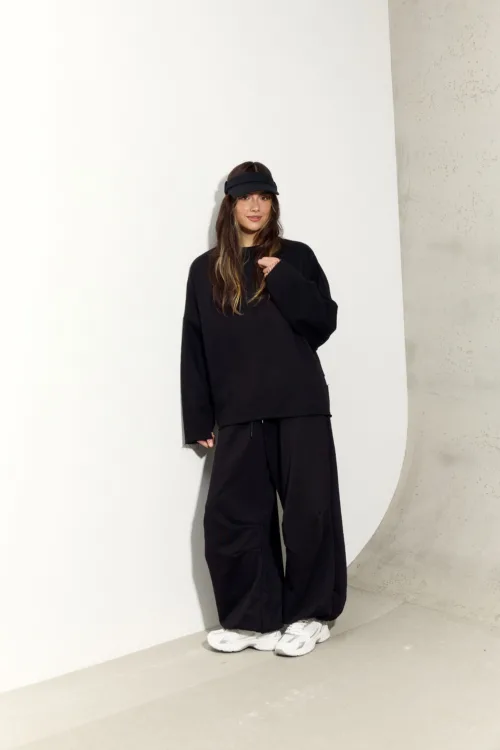 Woman in an oversized black sweatshirt and black wide-leg pants, wearing a black visor and white sneakers, standing against a white wall with a concrete edge.