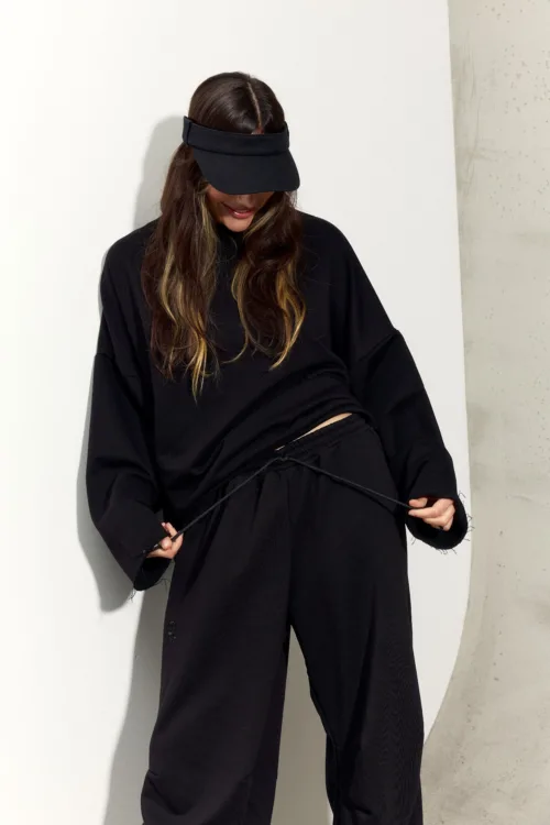 Person wearing an oversized black outfit with a black visor hat, standing against a light wall, adjusting a drawstring on their pants.